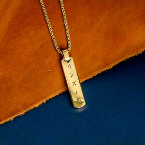May include: Gold chain necklace with a rectangular pendant engraved with Japanese characters. The pendant is on a brown leather surface.