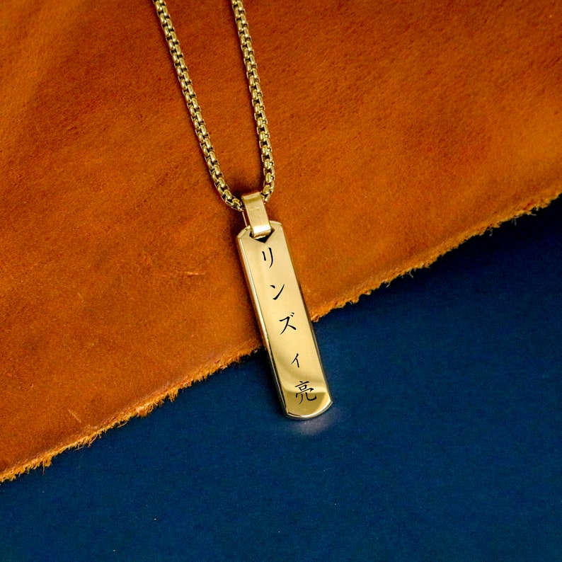 May include: Gold chain necklace with a rectangular pendant engraved with Japanese characters. The pendant is on a brown leather surface.