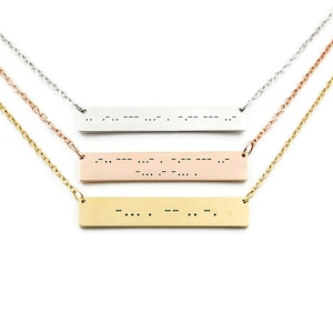 May include: Three silver, rose gold, and gold necklaces with bar pendants engraved with braille dots. The necklaces are layered on a white background.