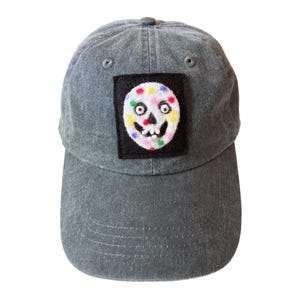 May include: A gray baseball cap with a curved brim. The cap features a black square patch with a colorful skull design. The skull has a white base with colorful polka dots and a smiling face.