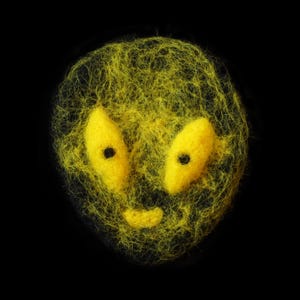 May include: A whimsical, handmade yellow felted face with two large, oval eyes and a small, curved mouth. The face is set against a stark black background, creating a striking contrast. The texture of the felted wool adds depth.