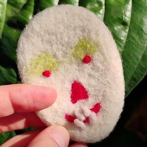 May include: A handmade, off-white felt skull-shaped brooch. The skull features red embroidered details for the mouth and eyes, and green and yellow accents for the eye sockets. The brooch is held by a hand against a green leaf background.
