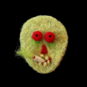 Peut inclure: Un visage de zombie en feutre fait à la main, avec des yeux rouges, un nez rouge et une cigarette dans la bouche. Le zombie a un teint vert clair et un fond noir.