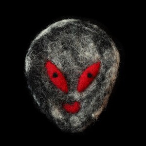 May include: A handmade felt alien face with red eyes and a red mouth. The alien is gray and white with a black background. The felt is textured and the alien has a slightly oval shape.