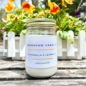 Op de afbeelding: Een witte kaars pot met een gouden deksel en een wit label met de tekst "CrenShaw Candles Citronella & Lavender Handcrafted". De kaars staat op een houten oppervlak voor een wit hek en een bed van oranje en paarse bloemen.