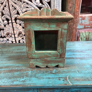 May include: A square wooden box with a distressed teal and brown finish, featuring a central glass panel on the front side, a scalloped top edge, and a matching scalloped base. The box has a sturdy frame construction with visible wood grain.