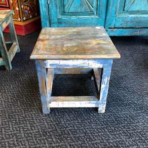 May include: A square wooden stool featuring a distressed finish with layers of blue, white, and natural wood tones. The stool has four sturdy legs connected by lower support rails, showcasing a weathered texture and visible wood grain throughout.