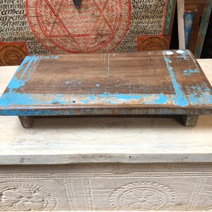 May include: A rectangular wooden riser with a natural brown finish and distressed bright blue paint along the edges. The low-profile platform is supported by four short, sturdy feet, showcasing a weathered texture and visible wood grain throughout.
