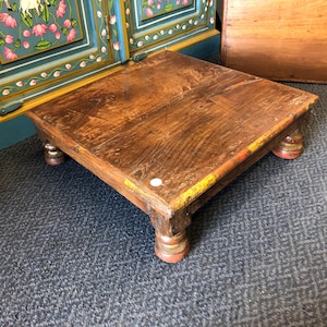 May include: A low, square wooden table with a dark brown, weathered finish and visible wood grain. The table features four short, turned legs with remnants of yellow and red paint, and a small white circular sticker on the top surface.