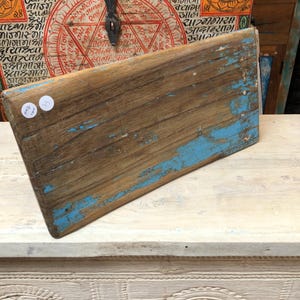 May include: A rectangular wooden board with a weathered finish, featuring patches of chipped blue paint over a natural brown wood grain. Two small white circular stickers are attached to the upper left corner of the surface, showing handwritten text.