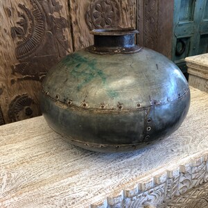 May include: A large, antique metal vase with a rounded body and a narrow neck. The vase is riveted together and has a weathered, grey-green patina. It sits on a white, carved wooden surface.