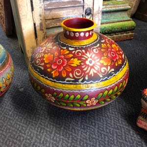 May include: A round metal pot featuring a deep red base colour with hand-painted floral patterns in orange, yellow, and green. The neck is painted yellow with white dots, and the body includes a horizontal yellow band with decorative green leaves.