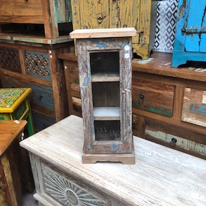 May include: A small, rustic wooden cabinet with a glass door and two shelves. The cabinet is painted in shades of brown, blue, and white, with a distressed finish. It is displayed on a white wooden surface, with other wooden furniture in the background.