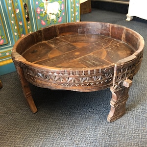 May include: A round, low-profile wooden table with a dark brown finish. The tabletop features a cross-shaped inlay pattern, while the exterior rim and the three sturdy, carved legs are decorated with intricate geometric, triangular, and linear carvings.