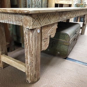 May include: A rectangular wooden bench with a weathered, light brown finish. The apron features carved geometric patterns, and the legs are thick and sturdy. A decorative carved wooden bracket is attached to the side of the frame under the top.