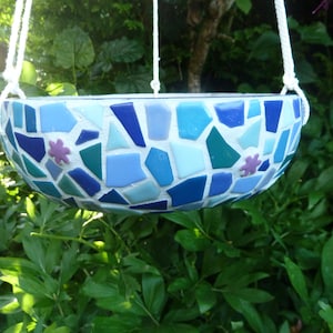 Könnte beinhalten: Ein hängender Mosaik-Pflanztopf mit blau-weißem Farbschema. Der Topf besteht aus kleinen, unregelmäßig geformten Fliesen in Blau-, Türkis- und Weißtönen, mit zwei kleinen lila Blumenakzenten. Er hängt an einem weißen Seil.