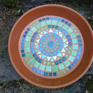Könnte beinhalten: Eine runde Untertasse aus Terrakotta mit einem kreisförmigen Mosaikdesign auf der Innenseite. Das Mosaik besteht aus kleinen, quadratischen und dreieckigen Glasfliesen in den Farben Limettengrün, Petrol, Dunkelblau und Weiß.