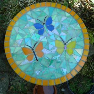 Könnte beinhalten: Ein runder Mosaik-Trittstein mit einem Rand aus orangefarbenen Fliesen. Die Mitte besteht aus irisierenden hellblauen und grünen Glasscherben mit drei Schmetterlingen in Blau, Orange und Gelb, die jeweils schwarze Drahtfühler haben.