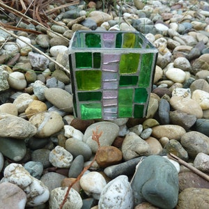 May include: A small, square, green and pink mosaic glass candle holder. The holder is made of small, square pieces of glass that are arranged in a geometric pattern. The holder is sitting on a bed of small, gray rocks.