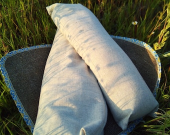 May include: Two light grey linen pillowcases are stacked on top of each other on a brown and blue woven mat. The mat is lying on a bed of green grass.