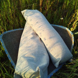 May include: Two light gray linen pillowcases are stacked on top of each other on a brown and blue woven mat. The mat is lying on a bed of green grass.