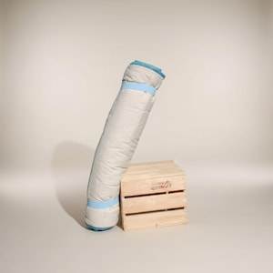 Pode incluir: Um saco-cama enrolado, de cor clara, com acabamentos e correias azuis, encostado a uma pequena caixa de madeira. O saco-cama parece ser feito de um material macio e acolchoado.