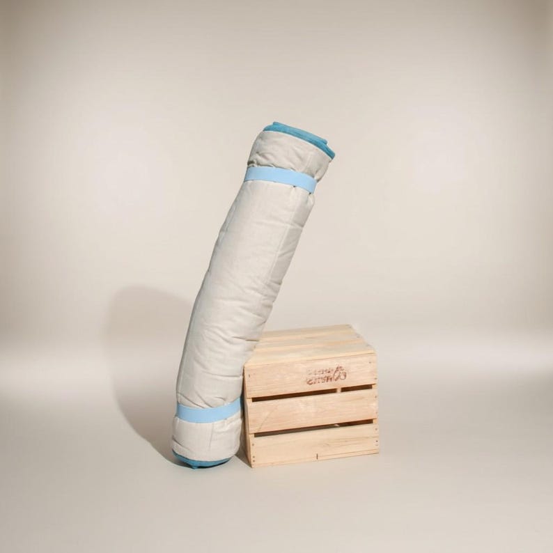 Pode incluir: Um saco-cama enrolado, de cor clara, com acabamentos e correias azuis, encostado a uma pequena caixa de madeira. O saco-cama parece ser feito de um material macio e acolchoado.