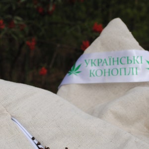 Puede incluir: Una funda de almohada blanca con una cremallera abierta, que revela c&aacute;scaras de trigo sarraceno marrones en el interior. La funda de almohada tiene una cinta blanca con texto verde que dice "українські коноплі" (C&aacute;&ntilde;amo ucraniano).