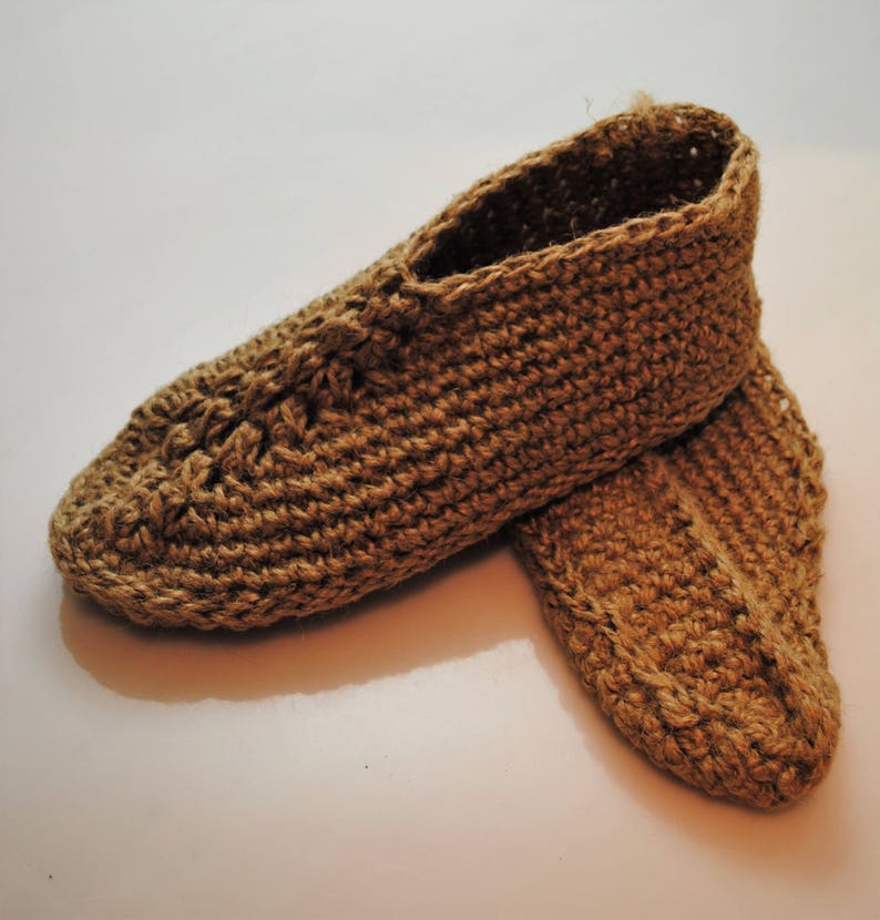 May include: A pair of handmade, brown crocheted slippers. The slippers are made of a thick, textured yarn, and feature a closed-toe design. The slippers are designed for indoor use and are a neutral color.