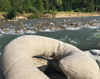 May include: A beige linen neck pillow resting on rocks by a river. The pillow is shaped like a crescent moon and has a textured surface.