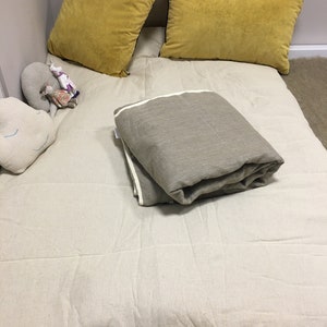 以下が含まれることがあります： 白いリネン製の布団マットレスに、マスタードイエローの枕が2つと、グレーのリネン製の毛布が折り畳んで置かれています。