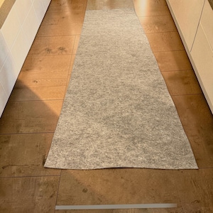 Puede incluir: Una alfombra larga y rectangular de fieltro gris claro colocada sobre un suelo de madera marrón. La alfombra está situada entre dos armarios blancos, creando un pasillo. El material de fieltro tiene un aspecto texturizado.