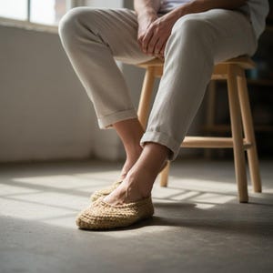 May include: A pair of beige, hand-crocheted slippers. The slippers are made of a textured yarn and have a rounded toe. The person is wearing light-colored pants and is sitting on a wooden stool.