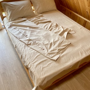 Op de afbeelding: Een beige linnen dekbedovertrek met een geknoopt overhemd erop gevouwen. Het dekbedovertrek ligt op een houten bedframe.