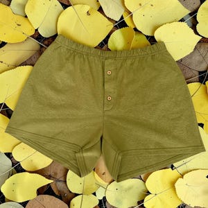 May include: Olive green boxer shorts with a button-up fly. The shorts are made of a soft fabric and have a comfortable elastic waistband. The background is a bed of yellow autumn leaves.