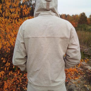 May include: A light beige hooded sweatshirt with a brown zipper pull. The sweatshirt is made of a soft, textured fabric. The person is standing in a woodland setting with autumn leaves.