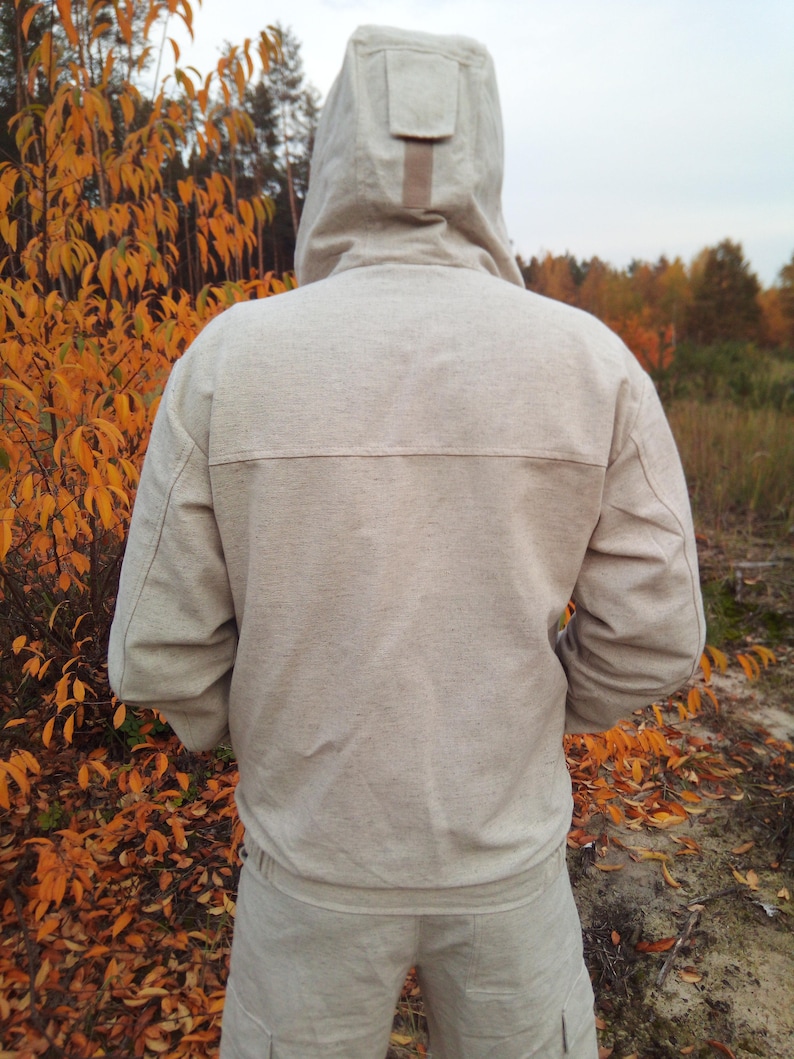 May include: A light beige hooded sweatshirt with a brown zipper pull. The sweatshirt is made of a soft, textured fabric. The person is standing in a woodland setting with autumn leaves.