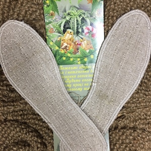 May include: Two beige, quilted insoles are displayed on a textured brown surface, with a Christmas-themed card in the background. The card features a cannabis plant, gifts, and festive decorations. The insoles are made of a natural fiber material.