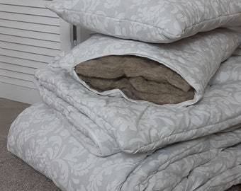 May include: A stack of light grey bedding with a white floral pattern, including pillows and a folded duvet. One pillow is open, showing a brown interior. The bedding is neatly arranged on a grey carpet, creating a cosy aesthetic.