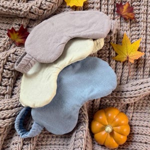 May include: Three sleep masks in neutral colors: lavender, cream, and blue. The masks are made of soft fabric and have elastic bands. The masks are arranged on a knitted blanket with autumn leaves and a small pumpkin.