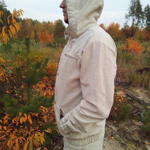 May include: A hooded jacket made of a light beige fabric. The jacket has a zipper closure and a drawstring hood. The jacket is worn by a person standing in a woodland setting.