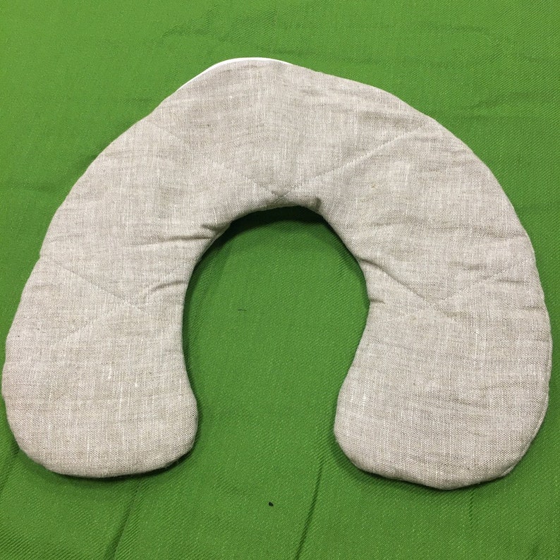May include: A beige linen neck pillow shaped like a horseshoe. The pillow is on a green surface.