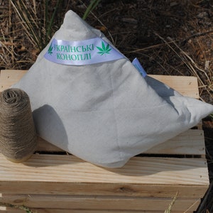Puede incluir: Una almohada blanca hecha de c&aacute;&ntilde;amo ucraniano, con una hoja de c&aacute;&ntilde;amo verde y el texto "Українські коноплі" en una cinta blanca. La almohada est&aacute; sentada en una caja de madera con un carrete de hilo de c&aacute;&ntilde;amo.