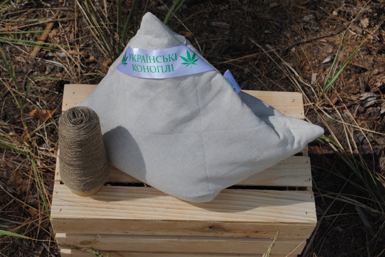 Puede incluir: Una almohada blanca hecha de c&aacute;&ntilde;amo ucraniano, con una hoja de c&aacute;&ntilde;amo verde y el texto "Українські коноплі" en una cinta blanca. La almohada est&aacute; sentada en una caja de madera con un carrete de hilo de c&aacute;&ntilde;amo.