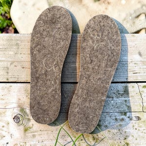 May include: A pair of brown felted wool insoles with a textured, fibrous surface. These shoe inserts feature a rounded heel and a tapered toe shape, designed to provide a layer of material inside footwear for added comfort and insulation.