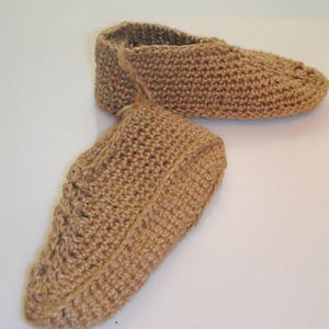 May include: A pair of handmade, brown crocheted slippers. The slippers are made from a natural fiber, possibly jute or hemp, and have a textured appearance. The design features a closed-toe style with a simple, comfortable shape. The slippers are shown on a white surface.
