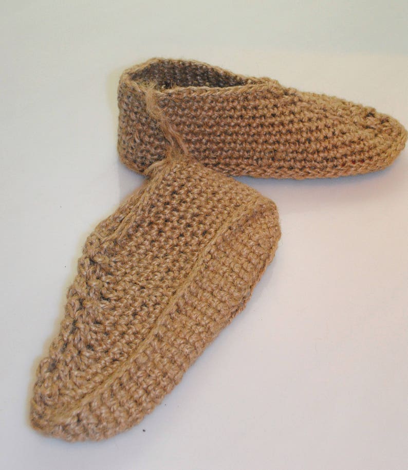 May include: A pair of handmade, brown crocheted slippers. The slippers are made from a natural fiber, possibly jute or hemp, and have a textured appearance. The design features a closed-toe style with a simple, comfortable shape. The slippers are shown on a white surface.
