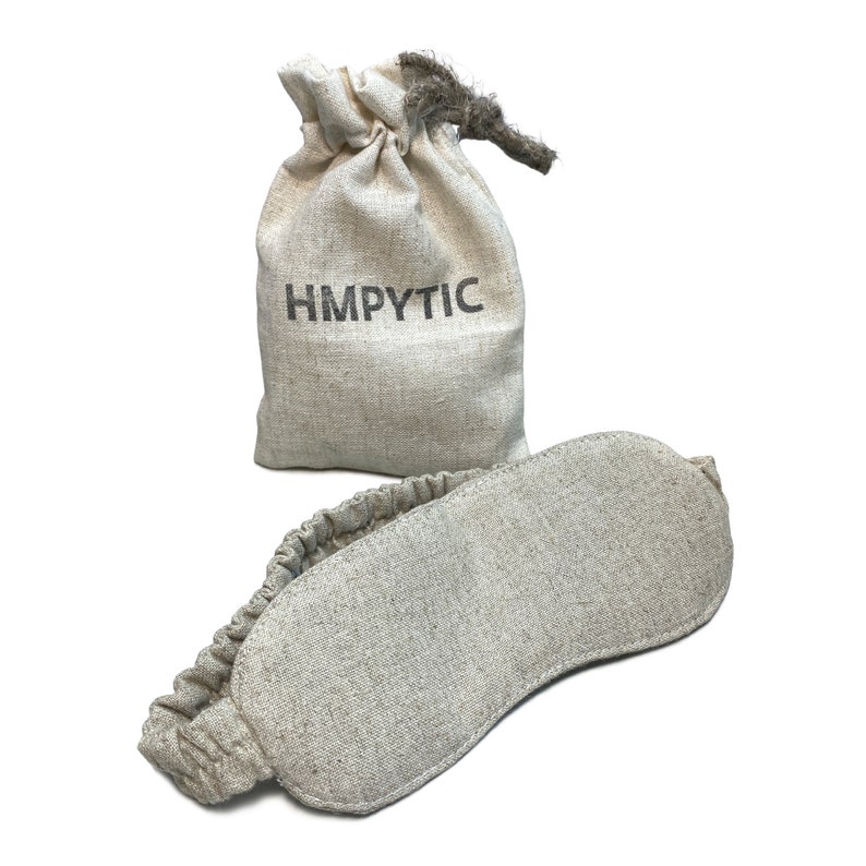 K&ouml;nnte beinhalten: Eine beige Schlafmaske mit Gummiband und passendem Kordelzugbeutel. Auf dem Beutel steht das Wort "HMPYTIC". Maske und Beutel sind aus einem strukturierten Stoff, wahrscheinlich Leinen oder einem &auml;hnlichen Material.