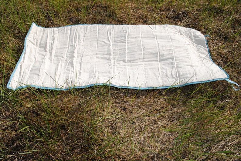 Pode incluir: Um tapete branco acolchoado com uma borda azul clara e uma al&ccedil;a em um canto. O tapete &eacute; retangular com costuras verticais e est&aacute; sobre grama seca.