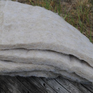 Pode incluir: Uma pilha de material fibroso bege claro, possivelmente isolamento ou estofamento, repousa sobre uma superf&iacute;cie de madeira envelhecida. O material parece macio e em camadas, com uma textura natural e org&acirc;nica. O fundo apresenta grama verde e folhagem.